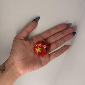 Puede incluir: Un objeto decorativo redondo rojo con una estrella amarilla y detalles en blanco y negro. El objeto se sostiene en una mano sobre un fondo blanco. Probablemente sea un pin o un bot&oacute;n.