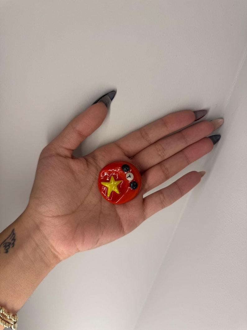 Puede incluir: Un objeto decorativo redondo rojo con una estrella amarilla y detalles en blanco y negro. El objeto se sostiene en una mano sobre un fondo blanco. Probablemente sea un pin o un bot&oacute;n.