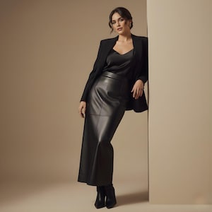May include: A woman wearing a black blazer, a black camisole, and a long black leather skirt. The outfit is paired with black ankle boots. The background is a neutral beige color.
