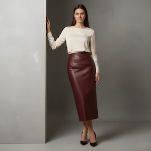 Op de afbeelding: Een vrouw draagt een crèmekleurige blouse met lange mouwen en een bordeauxrode leren potloodrok. De rok valt tot op de knieën. Ze draagt zwarte hoge hakken. De achtergrond is effen grijs.