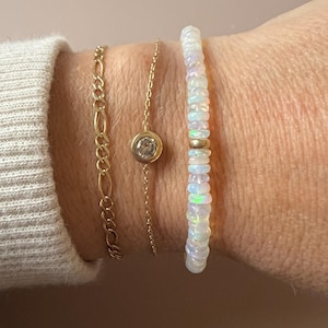 May include: Three gold bracelets are displayed. One is a chain link style, another is a delicate chain with a small, round pendant with a clear stone, and the third is a beaded bracelet with white opal beads and a gold spacer.
