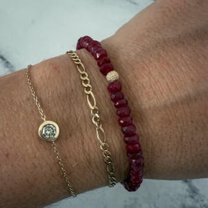 May include: Three bracelets on a wrist. A delicate gold chain bracelet with a bezel-set diamond, a gold chain bracelet with a Figaro link design, and a beaded bracelet with red gemstones and a gold bead.