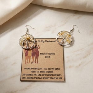 May include: Two silver-toned wire-wrapped tree earrings featuring yellow gemstone chips, displayed on a rectangular kraft paper card. The card text reads: Will You Be My Bridesmaid? MAID OF HONOR SOFIA. I FOUND MY MISTER, BUT I STILL NEED MY SISTER!