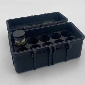 May include: A black, rectangular storage case with a hinged lid, open to reveal a series of circular compartments. A small glass bottle with a black cap sits in one of the compartments. The case has two latches on the front.