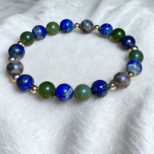 May include: A beaded bracelet featuring a mix of blue lapis lazuli, green jade, and gray-brown stone beads. The beads are separated by small gold-colored spacer beads, creating a colorful and elegant accessory.