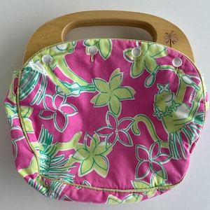 May include: A round fabric handbag featuring a vibrant pink background with a repeating pattern of light green monkeys and flowers. The bag has a light-colored wooden handle with a small palm tree icon etched into the right side and white buttons.