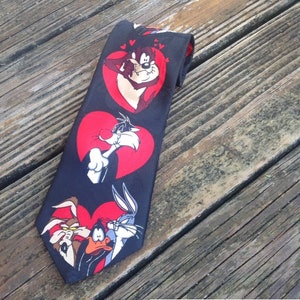 May include: A black silk tie with a repeating pattern of cartoon characters from the Looney Tunes series. The characters are Bugs Bunny, Daffy Duck, Sylvester the Cat, and Tweety Bird. Each character is depicted in a red heart.