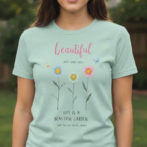 Op de afbeelding: Een lichtgroen t-shirt met het woord "beautiful" in roze script boven een bloemenontwerp. Het shirt heeft drie madeliefjes in blauw, wit en roze, met de tekst "Life is a beautiful garden" eronder. Vlinders zijn ook inbegrepen.