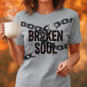 Könnte beinhalten: Graues T-Shirt mit dem Schriftzug "BROKEN SOUL" in einer verwaschenen Schriftart und einem Kettenmuster. Die Kette scheint an mehreren Stellen gebrochen zu sein. Das T-Shirt hat kurze Ärmel.