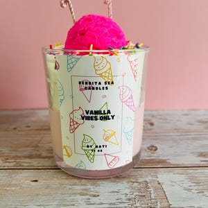 Puede incluir: Vela con aroma a vainilla en un tarro de cristal transparente, coronada con una bola de cera rosa brillante y chispas. El tarro presenta una etiqueta con ilustraciones de conos de helado y el texto "Vanilla Vibes Only".