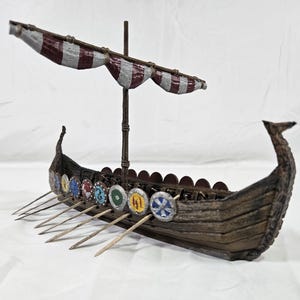 May include: A miniature wooden Viking longship model featuring a dark brown hull with a textured wood grain finish. The ship includes a central mast with a striped maroon and white sail, and a row of small, colorful circular shields along the side.