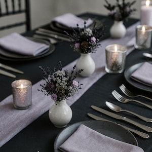 May include: A table setting with a black tablecloth, gray plates, and lavender napkins. Small white vases hold floral arrangements. Silverware and lit candles in glass holders complete the elegant dining scene.
