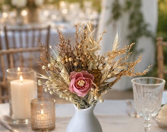 Op de afbeelding: Een kleine witte vaas met een boeket van gedroogde bloemen en een roze roos, op een tafel met kaarsen en glaswerk. Het arrangement bevat tarwe en grassen, waardoor een rustiek, natuurlijk middelpunt ontstaat.