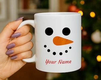 May include: White ceramic mug featuring a snowman face design with black eyes, a carrot nose, and a dotted smile. The text "Your Name" is printed in red at the bottom. The mug has a curved handle.