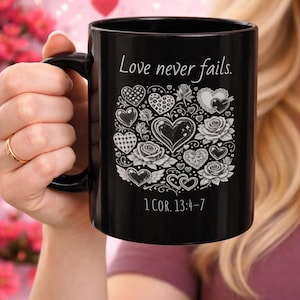 May include: Black ceramic mug with a handle, featuring the text "Love never fails." and the Bible verse 1 COR. 13:4-7. The design includes hearts and roses in white. The mug is held by a person.