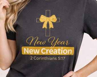 May include: Dark grey t-shirt with a gold cross and bow design. The text reads "New Year New Creation" and "2 Corinthians 5:17" in gold. A simple, faith-based design.