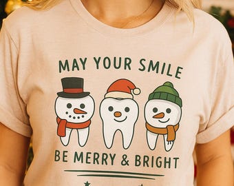 May include: Beige t-shirt with the text "MAY YOUR SMILE BE MERRY & BRIGHT" and three cartoon teeth dressed as snowmen. The snowmen teeth wear a top hat, Santa hat, and green beanie.