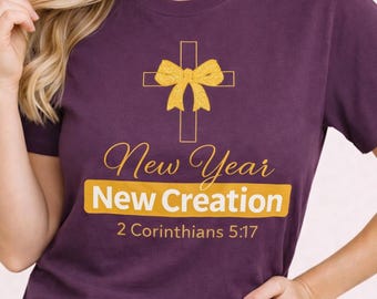 May include: A purple t-shirt featuring a gold cross and bow graphic. The text reads "New Year New Creation" in gold, with "2 Corinthians 5:17" underneath. The t-shirt has a crew neck.