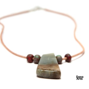 May include: A necklace with a light brown leather cord and a large, green, grey, and brown stone pendant. The pendant is surrounded by four small, red and green beads.
