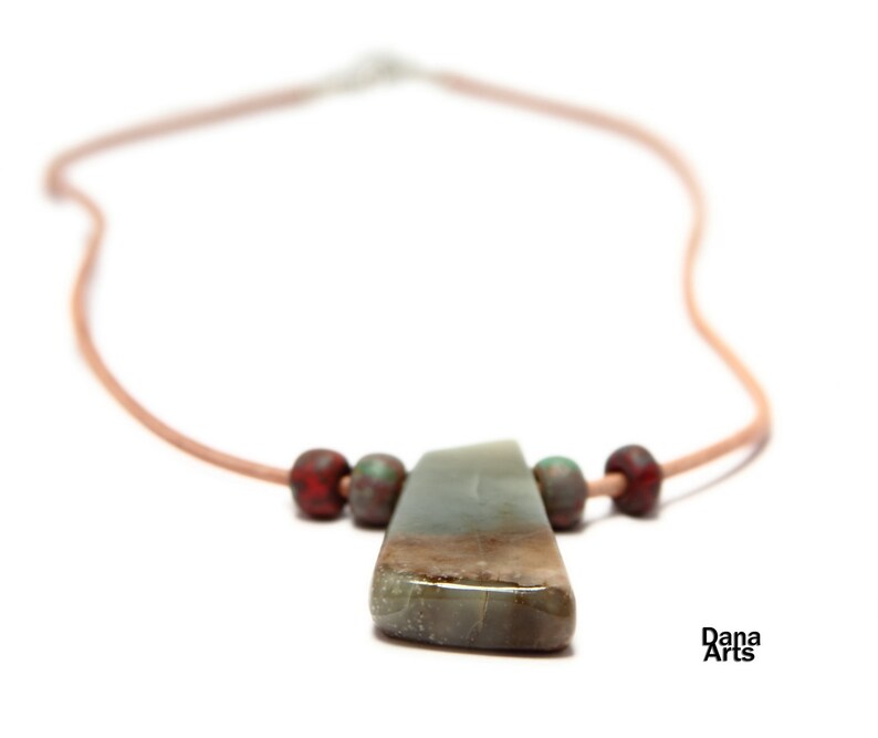 May include: A necklace with a light brown leather cord and a large, green, grey, and brown stone pendant. The pendant is surrounded by four small, red and green beads.