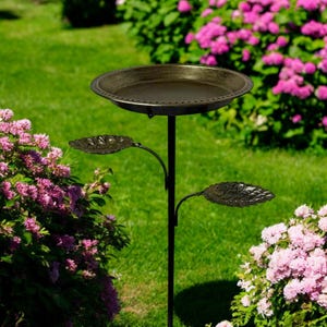 May include: A bronze bird bath with a decorative rim and a black metal stand. Two leaf-shaped accents extend from the stand. The bird bath is set in a garden with green grass and pink flowering bushes.