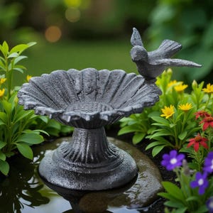 Puede incluir: Un bebedero para pájaros de hierro fundido gris oscuro con un cuenco decorativo en forma de girasol y una figura de pájaro posado. El bebedero está rodeado de flores coloridas y vegetación, creando un encantador acento de jardín.