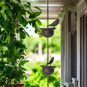 May include: A decorative rain chain featuring two bronze-coloured bird-shaped cups. The cups are suspended by a chain and designed to catch rainwater. The rain chain hangs from a porch roof, with green foliage in the background.