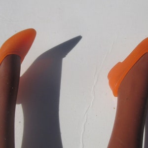 May include: A pair of orange plastic doll shoes. The shoes are on a pair of brown doll legs.