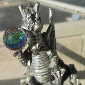 May include: A detailed silver-colored dragon figurine holding a colorful glass orb. The dragon has textured scales, wings, and a rocky base. The orb reflects a spectrum of colors, adding a magical element to the fantasy-themed sculpture.