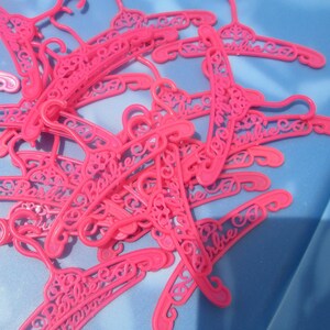 Op de afbeelding: Een stapel roze plastic hangers met het woord "Barbie" erop gedrukt.