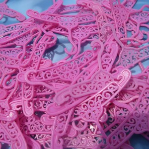 Op de afbeelding: Een stapel roze plastic poppenhangers met ingewikkelde ontwerpen. De hangers liggen allemaal door elkaar, sommige overlappen elkaar en sommige steken uit.