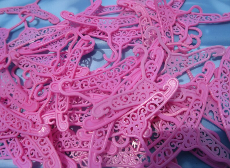 Op de afbeelding: Een stapel roze plastic poppenhangers met ingewikkelde ontwerpen. De hangers liggen allemaal door elkaar, sommige overlappen elkaar en sommige steken uit.