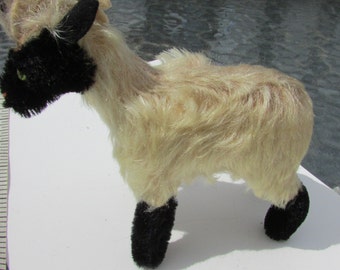 May include: A vintage toy ram with a cream-colored body, black face, ears, and legs, and a tan-colored curled horn. The ram has green eyes and a fluffy, textured appearance, standing on a white surface.
