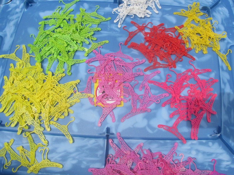 Op de afbeelding: Een grote verzameling plastic hangers in verschillende kleuren, waaronder geel, groen, roze en wit. De hangers bevinden zich allemaal in een plastic opbergdoos.