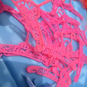 Op de afbeelding: Een stapel roze plastic hangers met het woord "Barbie" erop. De hangers zijn ontworpen met een decoratief, versierd patroon.