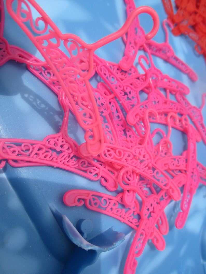 Op de afbeelding: Een stapel roze plastic hangers met het woord "Barbie" erop. De hangers zijn ontworpen met een decoratief, versierd patroon.