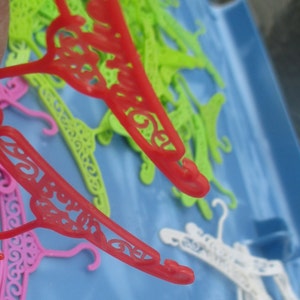 Op de afbeelding: Een close-up van een stapel kleurrijke plastic hangers. De hangers zijn in verschillende vormen en maten, waaronder sommige met decoratieve ontwerpen. De hangers liggen op een blauw oppervlak.