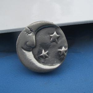 May include: A silver-toned metal charm featuring a crescent moon with a face and three stars.