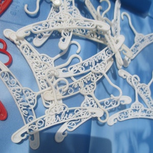 Op de afbeelding: Een stapel witte plastic hangers met het woord "Barbie" erop geschreven in een decoratief schrift.