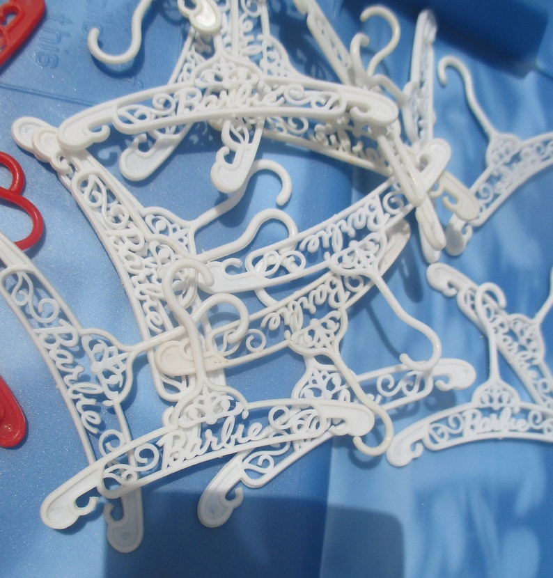 Op de afbeelding: Een stapel witte plastic hangers met het woord "Barbie" erop geschreven in een decoratief schrift.
