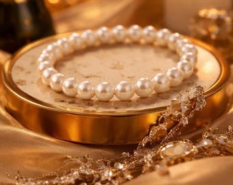 Könnte beinhalten: Ein Perlenarmband liegt auf einer goldgerahmten, marmorähnlichen Oberfläche. Das Armband besteht aus zahlreichen weißen Perlen. Die Szene umfasst eine brennende Kerze und goldfarbenen Stoff, was eine luxuriöse Ästhetik erzeugt.