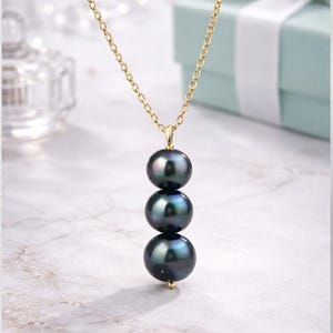 May include: A gold chain necklace with three dark, iridescent pearls. The pearls are stacked vertically, suspended from a gold pendant. The background includes a light blue gift box with a white ribbon.