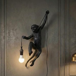 Op de afbeelding: Een zwarte aapvormige wandlamp met een gloeilamp in zijn hand. De lamp is bevestigd aan een grijze muur en een groene fluwelen fauteuil staat op de voorgrond. De lamp is een uniek decoratief item.