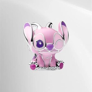 May include: A pink and purple character charm with silver accents. The charm features a cartoon character with large ears, one eye open and one eye closed. The charm is sitting down and has a silver base.