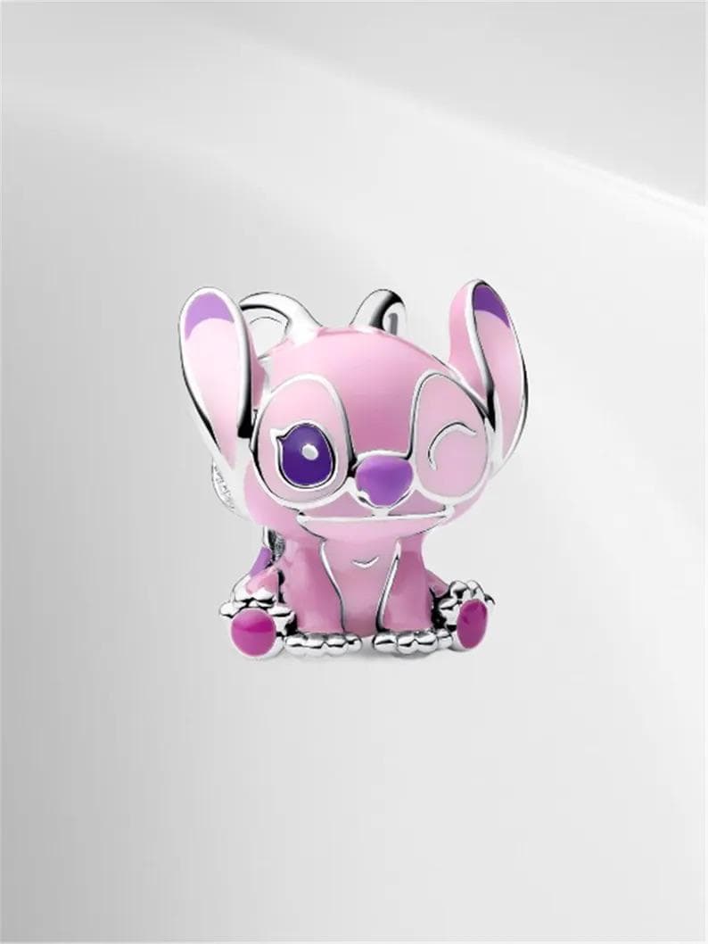 May include: A pink and purple character charm with silver accents. The charm features a cartoon character with large ears, one eye open and one eye closed. The charm is sitting down and has a silver base.