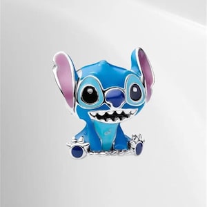 May include: A blue and silver charm featuring the character Stitch from the Disney movie Lilo & Stitch. The charm has large black eyes, a wide grin, and large ears with pink inner ears. The charm is made of metal and enamel.