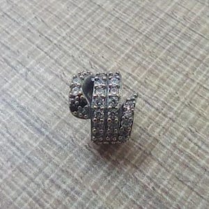 Può includere: Un orecchino a spirale color argento con più file di piccole pietre preziose trasparenti. L'orecchino è posto su una superficie strutturata marrone chiaro, che mette in risalto il suo design intricato.