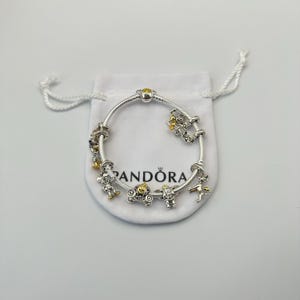 May include: A silver bracelet with gold and silver charms, including a carriage, a fairy, and a star. The bracelet is displayed on a white pouch with the brand name "Pandora" printed on it. The bracelet has a clasp with a yellow stone.