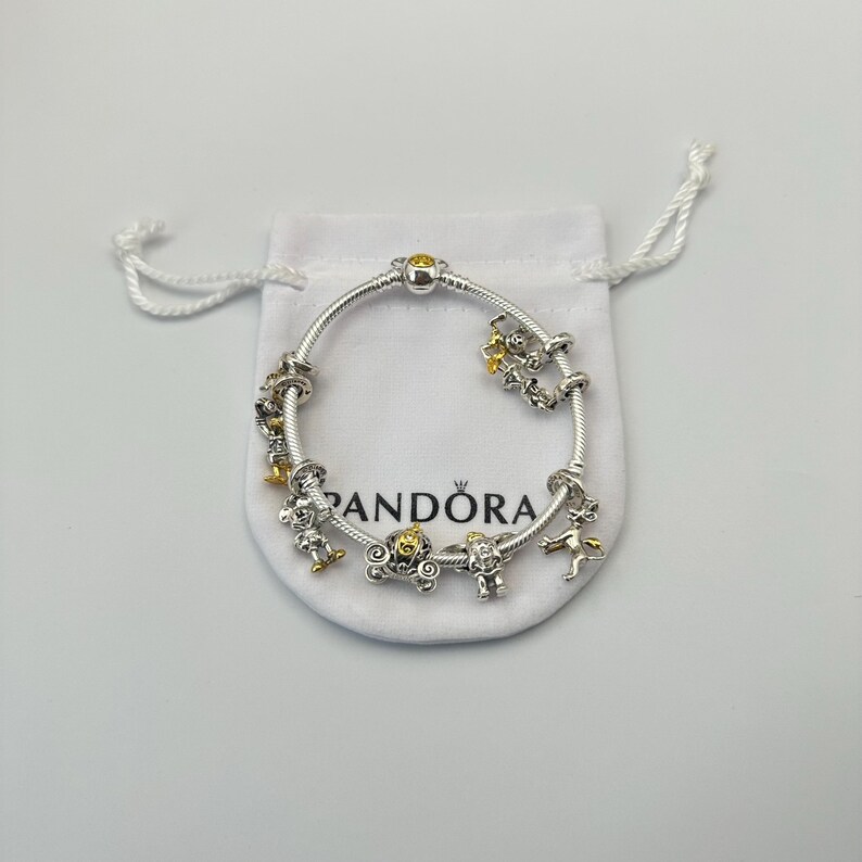 May include: A silver bracelet with gold and silver charms, including a carriage, a fairy, and a star. The bracelet is displayed on a white pouch with the brand name "Pandora" printed on it. The bracelet has a clasp with a yellow stone.