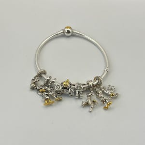 May include: Silver charm bracelet with multiple charms. Charms include a carriage, characters with gold accents, and other decorative elements. The bracelet is on a white background.